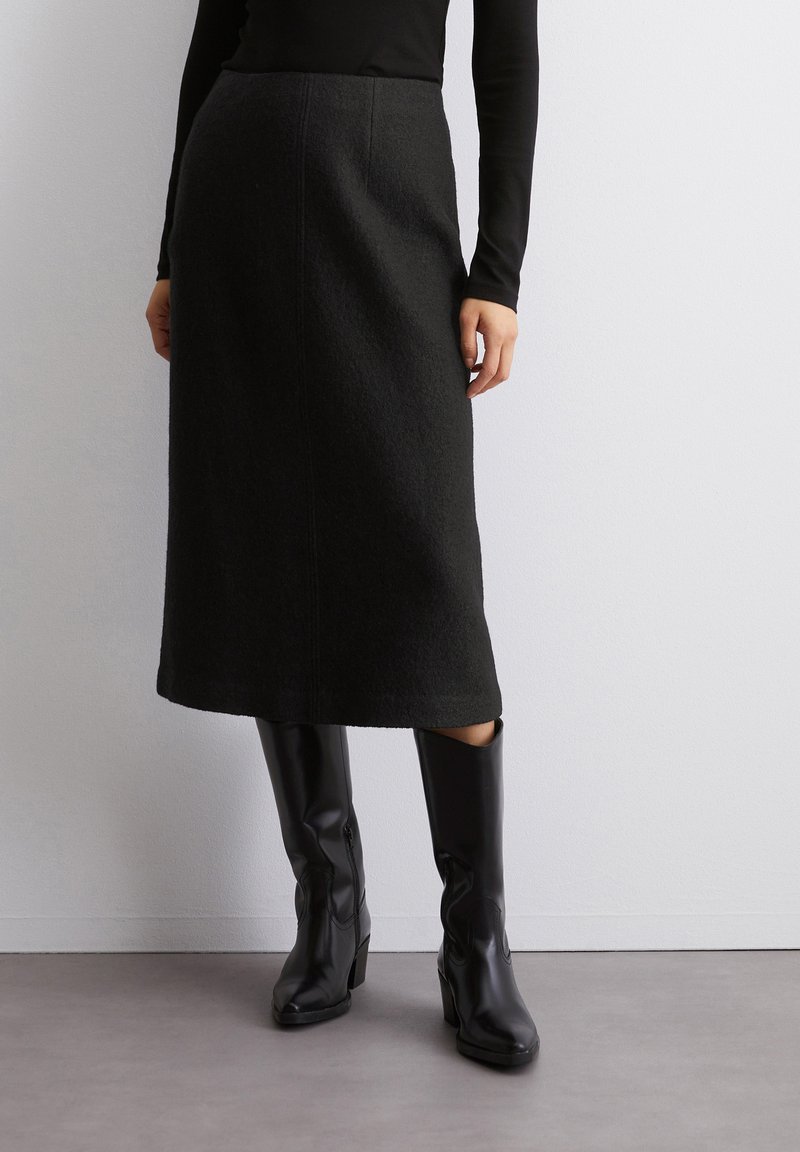 Schwarzer Woll-Midirock mit gerader Silhouette, sichtbaren Nahtdetails, kombiniert mit knielangen schwarzen Lederstiefeln. Strukturstoff.