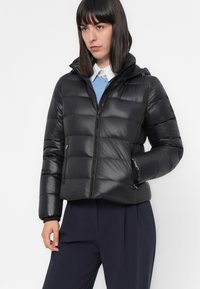 Vrouw in een zwarte gewatteerde pufferjack met capuchon, blauwe trui, witte overhemd en donkere broek voor een effen achtergrond.