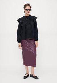 Svart teksturert bluse med volanger, kombinert med en tettsittende burgunder fuskeskinn midi-skjørt. Modellen har på seg svarte katteøye-solbriller og svarte sko.