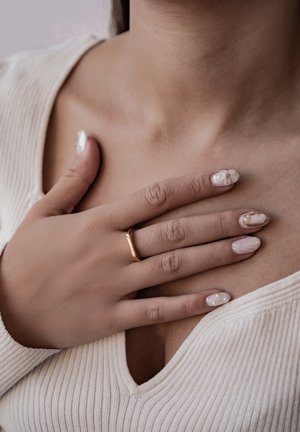 Goldring, der an einer Hand mit kunstvollem Nageldesign getragen wird; die Nägel zeigen weiße Akzente und florale Muster. Die Haut ist glatt, der Hals ist sichtbar.