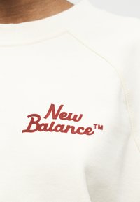 Κοντινό πλάνο λευκού φούτερ με κόκκινο κέντημα λογότυπου "New Balance™" στο στήθος κοντά στη ραφή του ώμου.