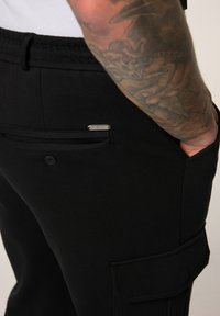 Zwarte cargobroek van gladde stof, met zijzakken, een elastische tailleband en een klein metalen logo-accent.