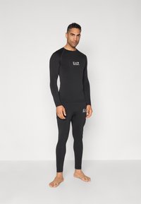 Juodas prigludęs termografinis komplektas su ilgomis rankovėmis ir legginsais, su EA7 logotipu ant krūtinės ir šlaunų, lygi tekstūra ir elastinga medžiaga.