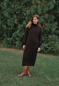 Brun strikket kjole med høj hals og lange ærmer, med knapper i siden. Kombineret med brune snørelange støvler og ribstrikkede sokker. Grøn baggrund med løv.