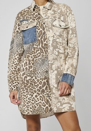 Long jacket with mixed animal print, digital camouflage, and denim patches, worn by a person with hands relaxed at sides.