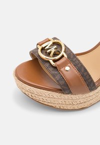 Gros plan sur une sandale compensée marron avec semelle tissée, brides en cuir et à motifs, et boucle dorée avec le logo MK sur la bride avant.