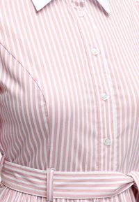 Κοντινή λήψη ενός ροζ και λευκού πουκάμισου-φόρεμα με κάθετες λωρίδες, λευκά κουμπιά και μια ζώνη με τις ίδιες ρίγες δεμένη στη μέση.