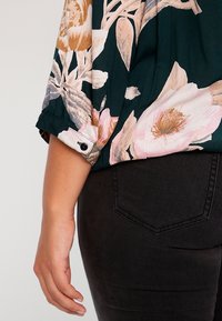 Bras et main d'une femme reposant à côté d'un pantalon noir avec une poche arrière, portant un chemisier foncé à motifs floraux roses, beige et blancs.