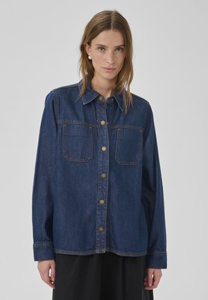 Nő sötétkék farmeringben, két mellzsebbel és aranyszínű gombokkal, sima szürke háttér előtt állva.