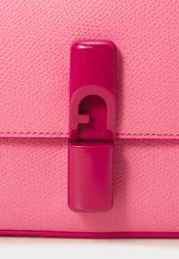 Pink textured leather handbag close-up featuring a glossy magenta clasp with a stylized logo cutout and contrasting stitching.