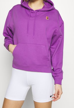 Un sweat à capuche violet avec une poche avant et un petit logo multicolore. Porté avec un short blanc arborant un logo Nike noir. Tissu doux.