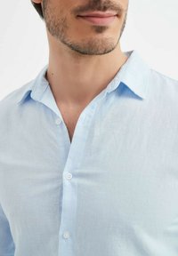 Chemise en lin bleu clair avec un col boutonné, présentant une texture subtile et deux boutons blancs visibles sur le devant.