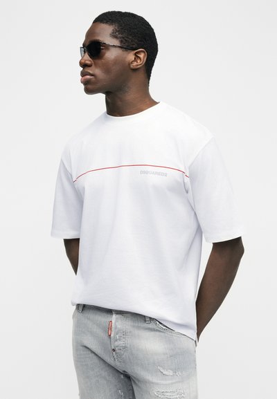 Άνδρας που φοράει μαύρα γυαλιά ηλίου, λευκό μπλουζάκι DSQUARED2 με κόκκινη γραμμή και ανοιχτόχρωμο τζιν με φθορές, στέκεται μπροστά σε ένα απλό λευκό φόντο.