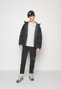 Giacca nera impermeabile con cappuccio, t-shirt bianca, pantaloni cargo neri e sneaker grigie. L'outfit presenta accenti del logo sugli indumenti.