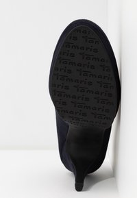 Chaussure à talon haut en daim bleu foncé avec un design effilé. La semelle extérieure noire présente un motif de logo en relief pour l'adhérence.