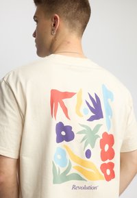 Naturlig beige bomulds t-shirt med et farverigt abstrakt design med blomsterformer i rød, gul, lilla og blå på ryggen, med tekst.