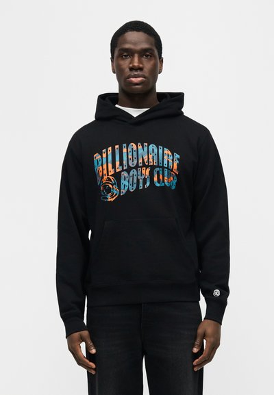 Νεαρός άνδρας με μαύρη μπλούζα με κουκούλα, με πολύχρωμο κείμενο "Billionaire Boys Club" και γραφικό κράνος αστροναύτη στο μπροστινό μέρος, στέκεται μπροστά σε απλό φόντο.