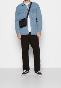 Camicia di denim azzurro chiaro, borsa a tracolla nera, t-shirt bianca con grafica, pantaloni neri con cuciture sulle ginocchia e sneakers Vans nere con dettagli bianchi.