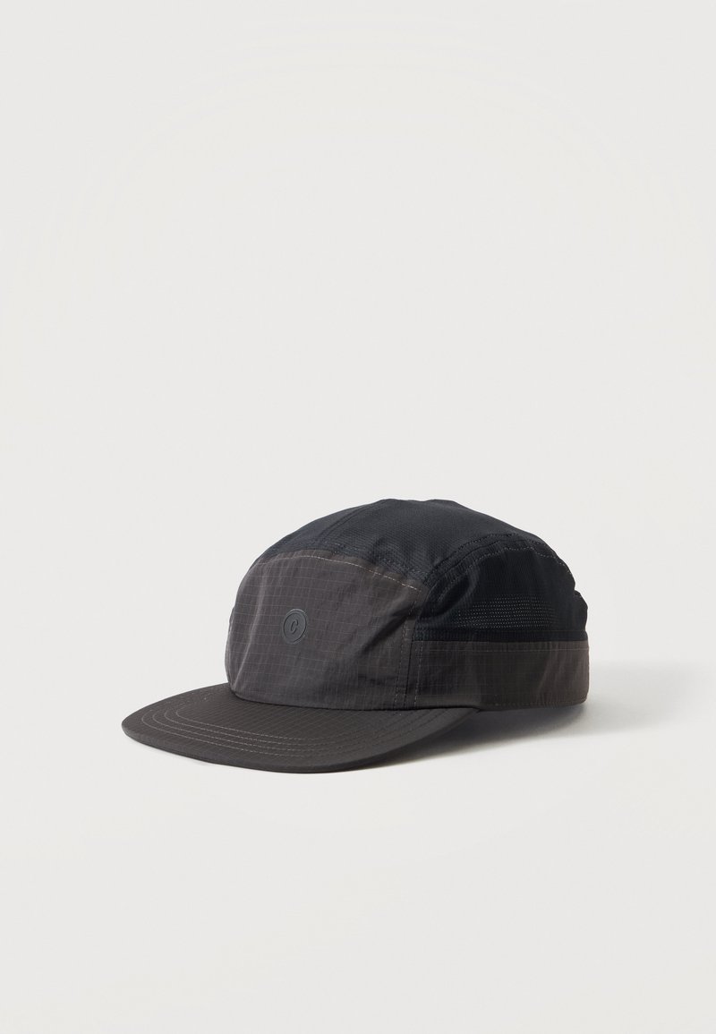 Casquette noire cinq panneaux avec une visière plate et un petit logo circulaire à l'avant, placée sur un fond blanc uni.