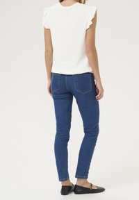 Kobieta ubrana w białą bezrękawnikową bluzkę z falbanami, niebieskie dopasowane dżinsy oraz czarne płaskie buty, stojąca tyłem do kamery na jednolitym tle.