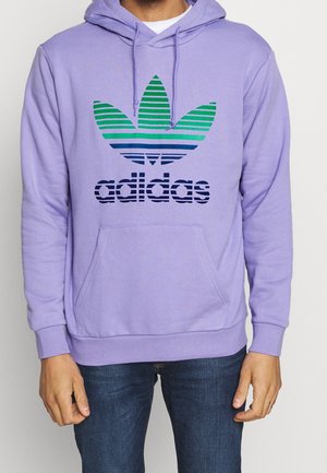 Persoon draagt een lavendelkleurige Adidas-hoodie met groen-blauw gestreept logo en donkerblauwe spijkerbroek, staand tegen een effen achtergrond.