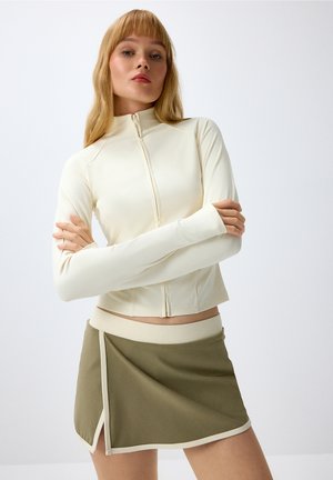 Woman with long blonde hair wearing a cream zip-up top and an olive green mini skirt with cream trim, standing with arms crossed.