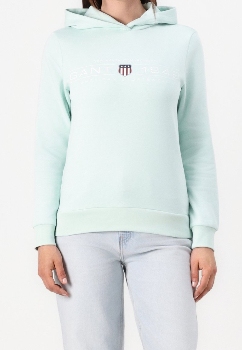 Personne portant un sweat à capuche vert menthe clair avec le logo GANT 1949 et un emblème en forme de bouclier, associé à un jean bleu clair sur fond blanc.