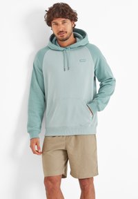 Man with short curly hair wearing seafoam green hoodie and beige shorts, standing against a white background with one hand in pocket.