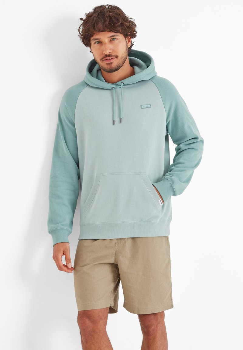 Man with short curly hair wearing seafoam green hoodie and beige shorts, standing against a white background with one hand in pocket.
