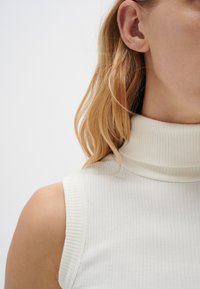 Ribbad vit ärmlös topp med hög polokrage, som har ett strukturerat tyg och en figurnära silhuett, som framhäver axel- och nackområdet.