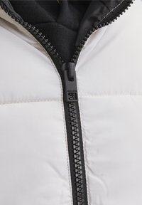 Biała pikowana kurtka puchowa z czarnym zamkiem błyskawicznym, z gładką powierzchnią i pikowaną fakturą. Wnętrze jest ciemne, prezentując warstwową konstrukcję.