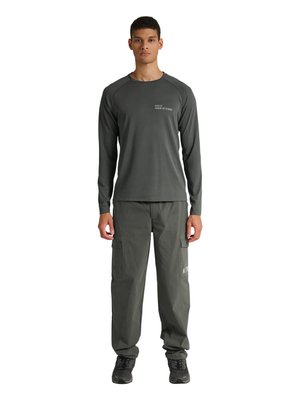 Mand stående vendt fremad iført mørkegrå langærmet skjorte med lille tekst og matchende cargobukser og sorte sneakers.