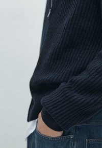 Maglione a coste in maglia blu navy scuro con cerniera, caratterizzato da tessuto strutturato e polsino aderente, abbinato a jeans azzurri.