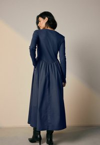 Navyblauwe jurk met een aansluitende bovenkant en een vloeiende geplooide rok, voorzien van lange mouwen en zijzakken. Draag met zwarte haklaarzen.