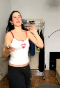 Frau trägt ein weißes Tanktop und tanzt in einem Raum mit einem Metallkleiderständer und einem beigen Sofa.