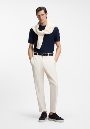 Man draagt marineblauw shirt met korte mouwen, crème broek, zwarte sneakers, met crème trui over de schouders gedrapeerd, staat met handen in de zakken.