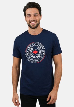 Homme souriant portant un t-shirt bleu marine avec l’inscription « Lee Cooper Originals East 1908 » et un logo de couronne de laurier imprimé sur le devant.