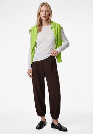Kvinde med blondt hår iført en hvid langærmet skjorte, brune løse bukser, sorte loafers og en knaldgrøn sweater, der er lagt over skuldrene.
