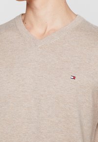 Beige V-Ausschnitt-Pullover aus weichem Stoff, mit einem kleinen gestickten Logo in Rot, Weiß und Blau, sowie gerippten Bündchen und einem gerippten Halsausschnitt.