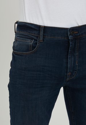 Dunkelblaue Denim-Jeans, getragen mit einem weißen Hemd, das hineingesteckt ist, und zeigt Vordertaschen, Gürtelschlaufen, Knopf- und Reißverschlussverschluss.