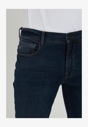 Jeans de mezclilla azul oscuro usados con una camisa blanca metida, mostrando los bolsillos frontales, las presillas del cinturón, el botón y la cremallera.