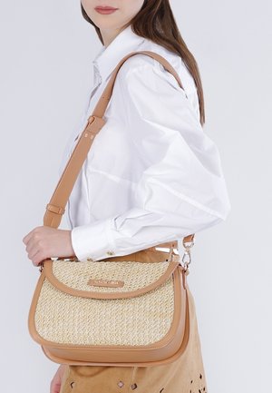 Mujer con camisa blanca que lleva un bolso tejido beige con ribete de cuero y correa ajustable para el hombro colgado sobre un hombro.