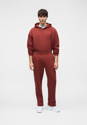 Młody mężczyzna w brązowej bluzie z kapturem i pasujących spodniach dresowych oraz białych trampkach, stojący na jednolitym jasnym tle.