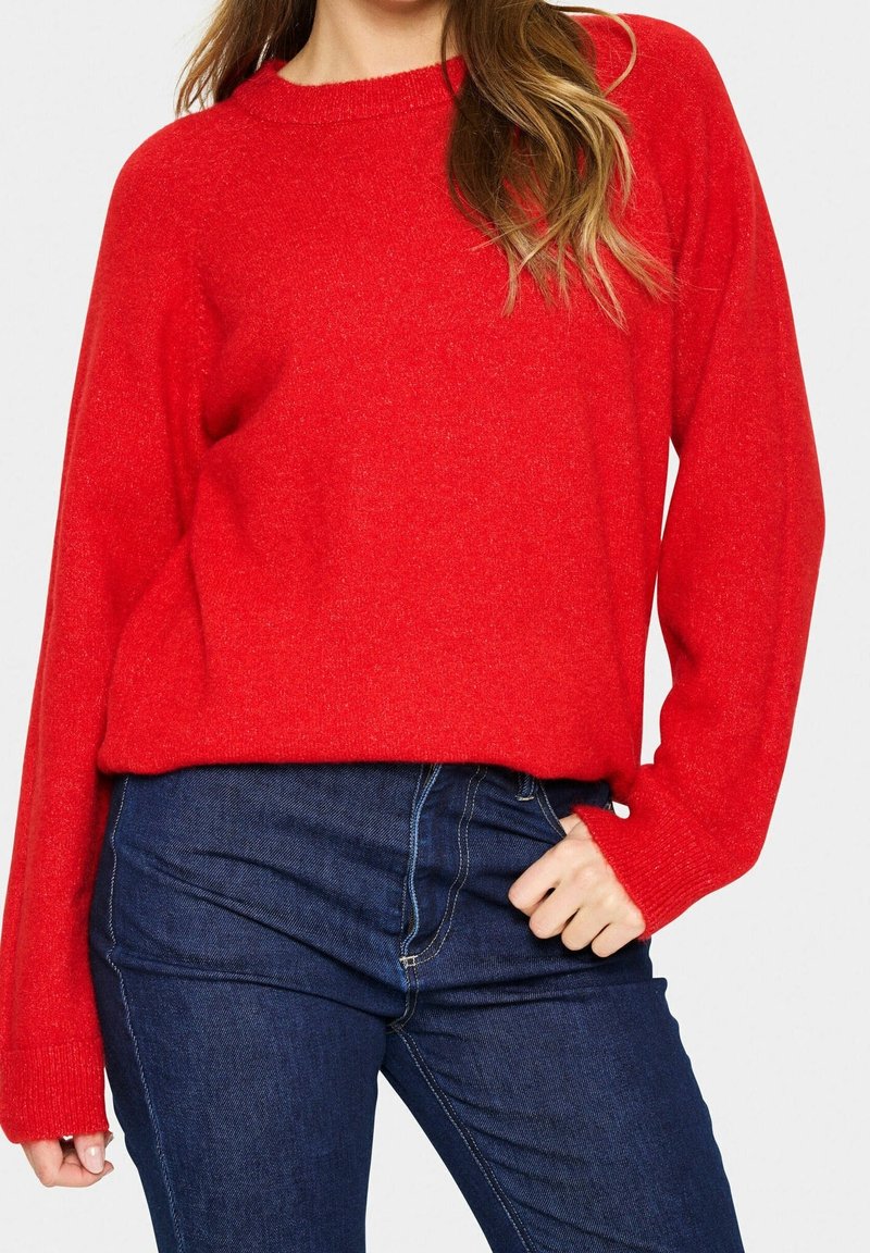Femme portant un pull rouge vif à manches longues associé à un jean en denim bleu foncé, posant avec une main près de la hanche.