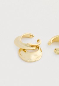 Un par de pendientes de aro abiertos de metal dorado brillante con un acabado martillado, de forma ligeramente irregular y redondeada, sobre un fondo blanco.