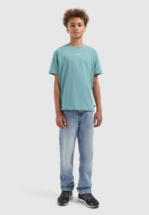 Jongeman met krullend haar, gekleed in een tealgroene t-shirt, lichtblauwe jeans en zwarte sneakers, staand tegen een effen lichte achtergrond.