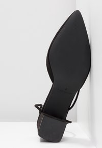 Chaussure noire à bout pointu avec un petit talon bloc, semelle tournée vers l'avant, reposant contre une surface d'angle blanche.