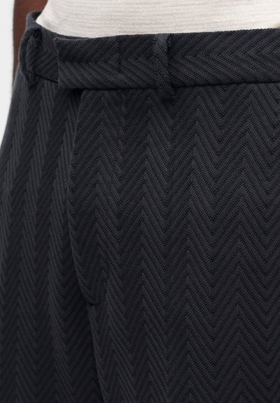 Μαύροι υφασμάτινοι παντελόνια με υφή και σχέδιο ψαροκόκαλο. Διαθέτουν θηλιά για ζώνη και πιέτες, που αναδεικνύουν ένα ραμμένο σχέδιο με δομημένο σχήμα.