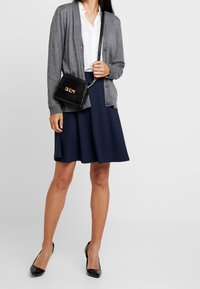 Mujer vestida con un cárdigan gris, blusa blanca, falda de tablas azul marino, tacones altos negros, y llevando un bolso de hombro texturizado negro.