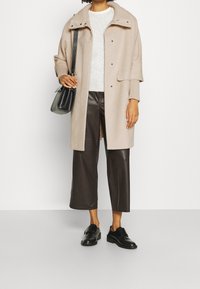 Beige, wollen blend jas met ribgebreide kraag, grijze trui, bruine leren cropped broek en zwarte schoenen; een zwarte handtas vasthoudend.